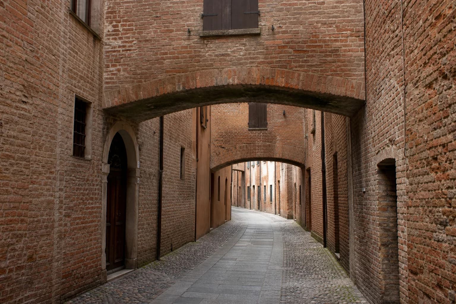 A narrow red-brick lane in Ferrara with covered medieval passages bridging Via delle Volte