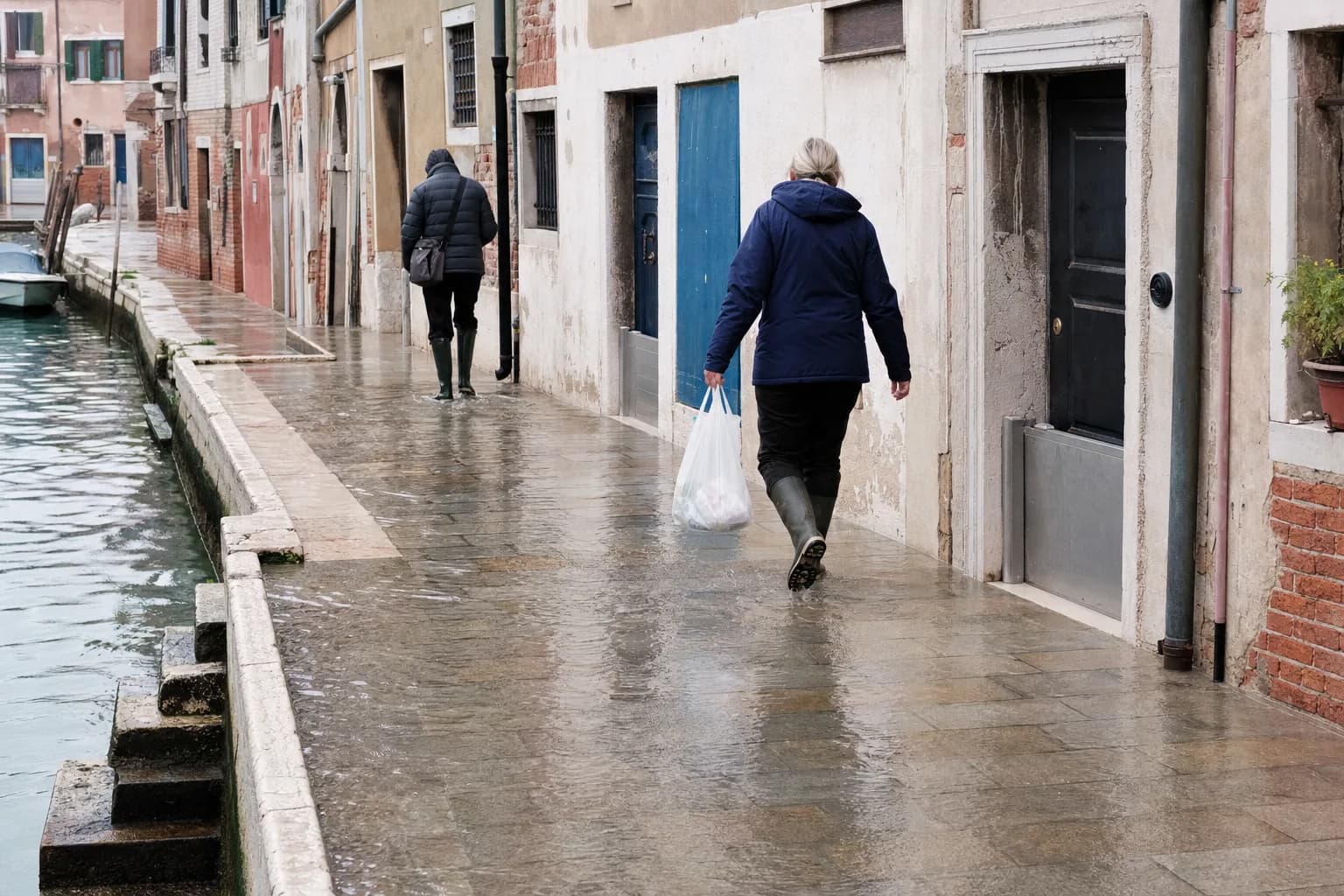 Two residents in waterproof boots walking along a shallowly flooded Venetian canal pavement